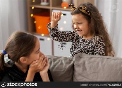 halloween, holiday and childhood concept - smiling little girls in party costumes playing with toy spider at home. girls in halloween costumes playing with spider