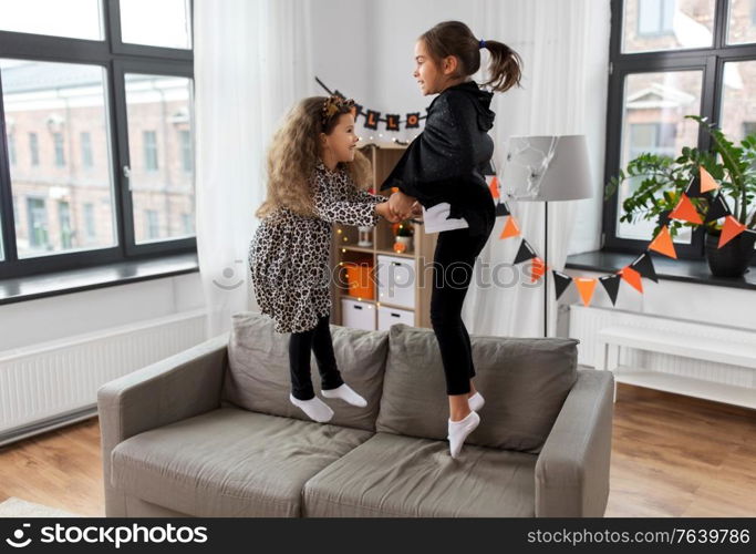 halloween, holiday and childhood concept - smiling little girls in party costumes jumping on sofa at home. girls in halloween costumes jumping on sofa