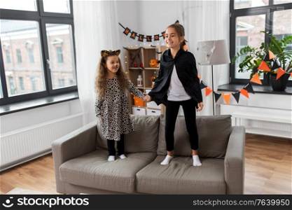 halloween, holiday and childhood concept - smiling little girls in party costumes jumping on sofa at home. girls in halloween costumes jumping on sofa