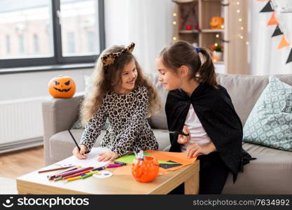 halloween, holiday and childhood concept - smiling little girls in party costumes doing crafts at home. girls in halloween costumes doing crafts at home