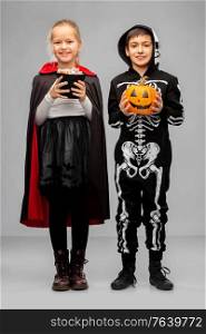 halloween, holiday and trick-or-treating concept - happy smiling boy and girl in costumes of dracula and skeleton with candies and jack-o-lantern pumpkin over grey background. happy children in halloween costumes with candies