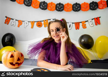 Halloween holiday concept. Cute little girl in witch costume sitting behind a table in Halloween theme decorated room. Halloween with safety measures from Covid-19. Halloween holiday concept. Cute little girl in witch costume sitting behind a table in Halloween theme decorated room.