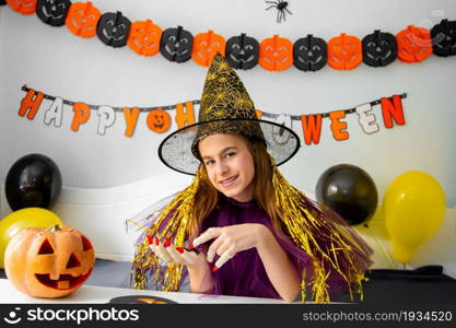 Halloween holiday concept. Cute little girl in witch costume sitting behind a table in Halloween theme decorated room. Halloween with safety measures from Covid-19. Halloween holiday concept. Cute little girl in witch costume sitting behind a table in Halloween theme decorated room.