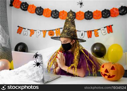 Halloween holiday concept. Cute little girl in witch costume sitting behind a table in Halloween theme decorated room. Halloween with safety measures from Covid-19. Halloween holiday concept. Cute little girl in witch costume sitting behind a table in Halloween theme decorated room.