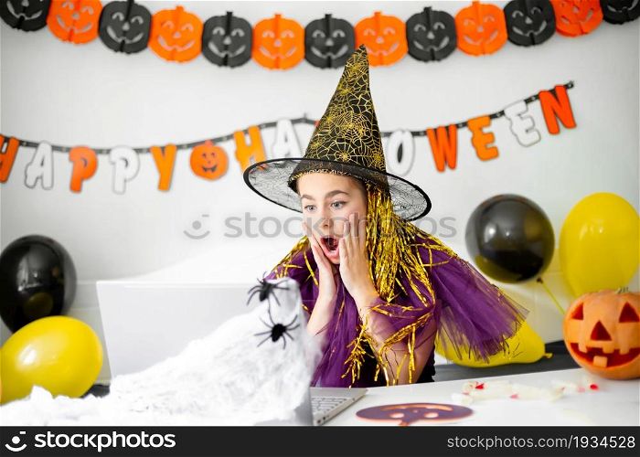Halloween holiday concept. Cute little girl in witch costume sitting behind a table in Halloween theme decorated room. Halloween with safety measures from Covid-19. Halloween holiday concept. Cute little girl in witch costume sitting behind a table in Halloween theme decorated room.
