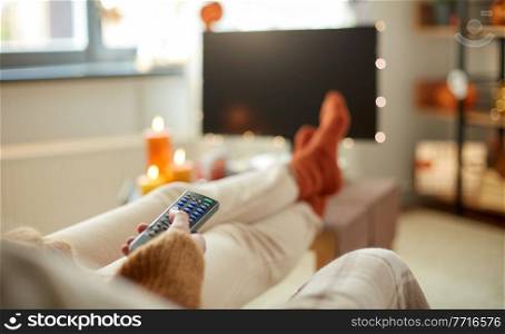halloween, holidays and leisure concept - close up of young womanwith remote control watching tv and resting her feet on table at cozy home. young woman watching tv at home on halloween