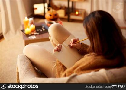 halloween, holidays and leisure concept - young woman with pencil writing to diary and resting her feet on table at cozy home. woman writing to diary at home on halloween