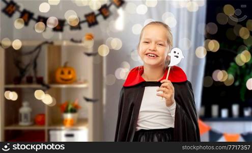 halloween party, holiday and childhood concept - happy smiling girl in black dracula costume with cape and ghost over lights and decorated home room background. girl in costume of dracula with cape on halloween