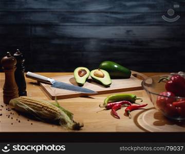 Halved and whole ripe avocados on wooden board with knife. Low key shot, light on board, some vegetables around on table. Copy space.