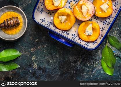 Halves of peaches with cheese and honey in baking dish on dark rustic background, top view. Baked peaches preparation.