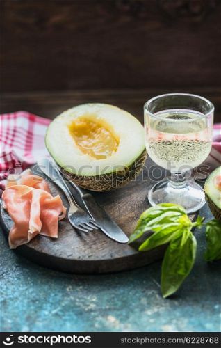 Ham and melon half with white sparkling wine on cutting board on rustic kitchen table, place for text. Italian food concept