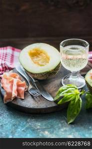 Ham and melon half with white sparkling wine on cutting board on rustic kitchen table, place for text. Italian food concept