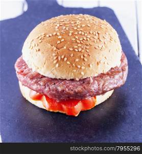 Hamburger sandwich with tomato over black stone chopping board, square image