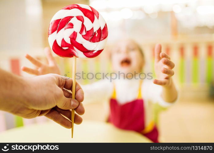 Hand gives handmade lollipop to happy little girl. Children in workshop at pastry shop. Holiday fun in candy store. Fresh cooked sugar caramel