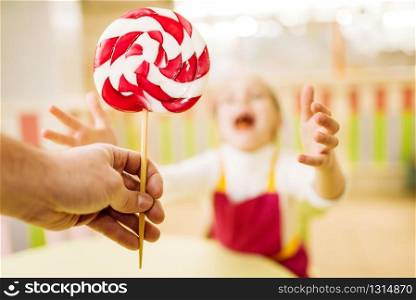 Hand gives handmade lollipop to happy little girl. Children in workshop at pastry shop. Holiday fun in candy store. Fresh cooked sugar caramel