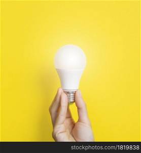 Hand holding a LED light bulb on bright yellow background. Using economical and environmentally friendly light bulb concept. Idea concept. Energy saving lamp in woman&rsquo;s hand.. Hand holding a LED light bulb on bright yellow background. Using economical and environmentally friendly light bulb concept. Idea concept. Energy saving lamp in woman&rsquo;s hand