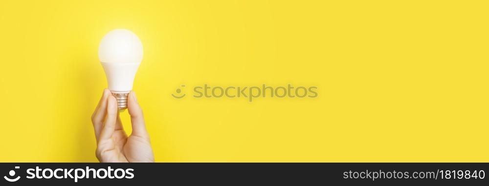 Hand holding a LED light bulb on bright yellow background. Using economical and environmentally friendly light bulb concept. Idea concept. Energy saving lamp in hand. Long Banner with copy space.. Hand holding a LED light bulb on bright yellow background. Using economical and environmentally friendly light bulb concept. Idea concept. Energy saving lamp in hand. Long Banner with copy space