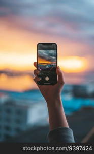 Hand holding a smartphone taking a photograph of the sea at sunset