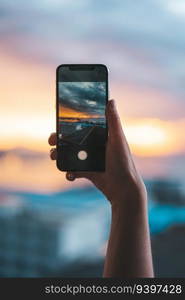 Hand holding a smartphone taking a photograph of the sea at sunset