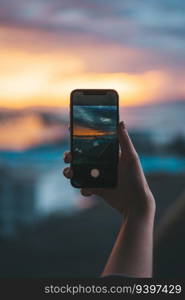 Hand holding a smartphone taking a photograph of the sea at sunset