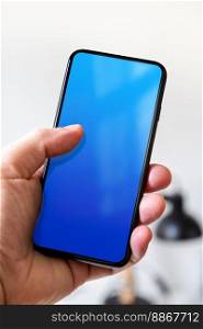 Hand holding a smartphone with blank blue screen. White office background.. Hand holding a smartphone with blank blue screen. Office background.