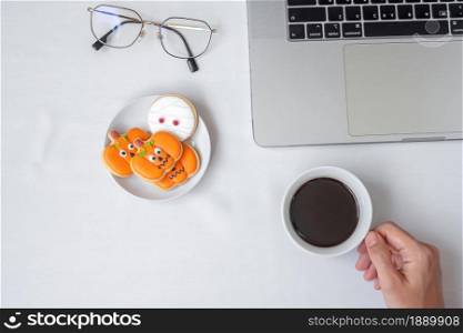Hand holding coffee cup and eating Halloween Cookies during using computer laptop. Happy Halloween, online shopping, Hello October, fall autumn, Festive, party and holiday concept