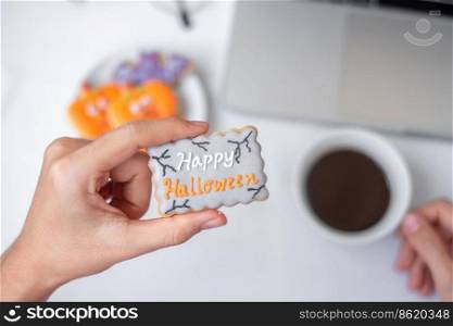 Hand holding Halloween Cookie and drinking coffee during using computer laptop. Happy Halloween, online shopping, Hello October, fall autumn, Festive, party and holiday concept