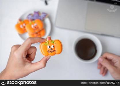 Hand holding Halloween pumpkin Cookie and drinking coffee during using computer laptop. Happy Halloween, online shopping, Hello October, fall autumn, Festive, party and holiday concept