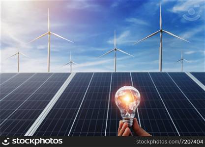 hand holding lightbulb with solar panel and wind turbine background. clean energy in nature concept