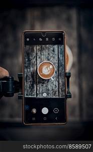 Hand holding mobile smart phone or cell telephone digital camera taking photo to Cup of Hot Cappuccino coffee with fresh milk . Hand holding mobile smart phone or cell telephone digital camera taking photo to Cup of Hot Cappuccino coffee