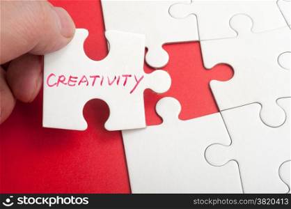 Hand holding puzzle piece which written creativity word and inserting it into group of white paper jigsaw puzzles