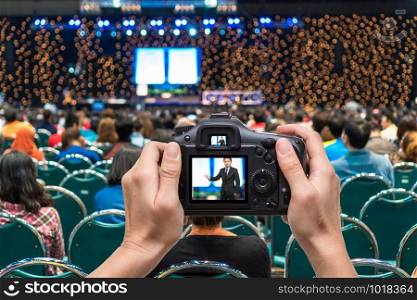Hand holding the camera taking photograph to the businessman on the stage over the Rear view of Audience in the conference hall in Exhibition Center background, Business meeting and education concept
