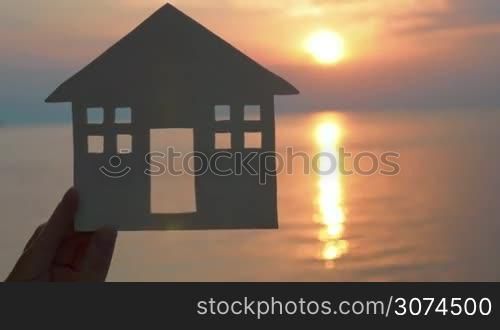 Hand holding white cut paper house against of sea at sunset. Unrecognizable.