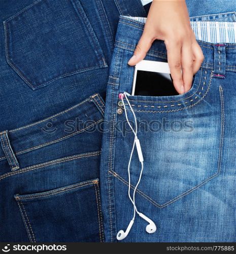 hand holding white smartphone with a blank black screen and headphones is in the front pocket of blue jeans, close up