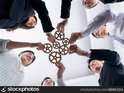 Hand holding wooden gear by businesspeople wearing suit for harmony synergy in office workplace. Bottom view office worker hand make chain of gear into collective unity form.. Group of people making chain of gears into collective form for harmony symbol.