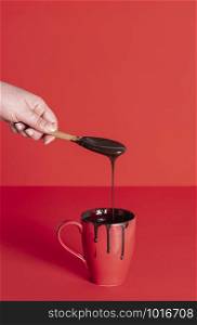Hand holding wooden spoon with dripping melted chocolate into red mug. Cup of chocolate glaze. Pouring melted chocolate. Christmas dessert decorating