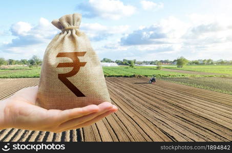 Hand holds out a indian rupee a farm field. Financial support, subsidies for agriculture. Farm loans, economic preferences. Purchase or lease rent of land. Payment of taxes for use natural resources.