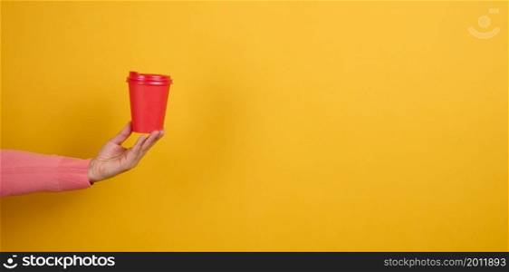 hand holds paper cardboard red cup for coffee, yellow background. Eco-friendly tableware, copy space