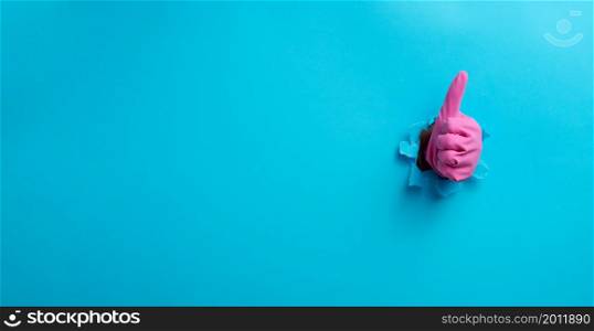 hand in a pink household rubber glove sticks out of the torn hole and shows a gesture okay, thumb up. Blue background