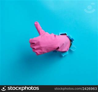  hand in a pink household rubber glove sticks out of the torn hole and shows a gesture okay, thumb up. Blue background