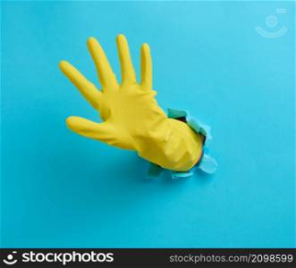hand in a yellow latex cleaning glove sticks out of a torn hole in a blue paper background
