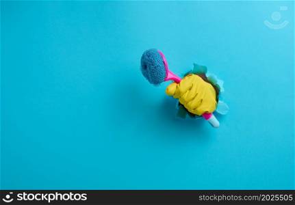 hand in a yellow rubber glove holds a plastic cleaning brush on a blue background, copy space