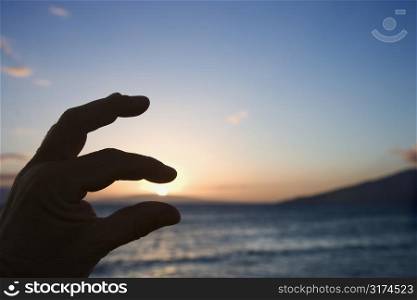 Hand in foreground with fingers around sunrise over the coast of Kihei, Maui, Hawaii, USA.