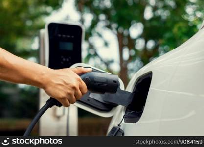 Hand inserting EV charging plug to electric vehicle in focus shot with blurred background of outdoor natural greenery. Progressive sustainable energy powered electric charging station for rechargeable. Focus hand insert progressive EV charger plug to EV car with blur background.