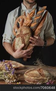Hand of a baker holding fresh organic bread and baguette on a dark background wooden table with dry flowers and round bread. Variety of bread hold men’s hands
