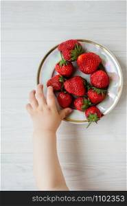 Hand of a child with a strawberry on a rustic background, a plate of strawberries. The concept of summer healthy eating. View from above, apartment.. Hand of a child with a strawberry on a rustic background, a plate of strawberries.