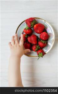 Hand of a child with a strawberry on a rustic background, a plate of strawberries. The concept of summer healthy eating. View from above, apartment.. Hand of a child with a strawberry on a rustic background, a plate of strawberries.