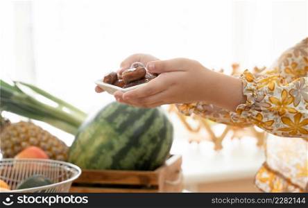 Hand of Arab muslim kid girl wearing hijab holding a plate full of sweet dry dates fruit on iftar time in ramadan kareem in kitchen at home with watermelon, pineapple, apples and vegetables