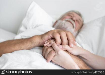 hand of senior man with smiling nurse, takes care and cheer on bed at nursing home