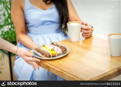 Hand of waiter puts a plate with eclairs on the wooden table for young woman drinking coffee in a cafe. Waiter puts plate with eclairs